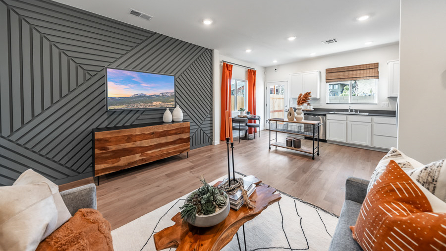 Open living space in Phoenix's model home, with an accent wall with carpentry details behind the wall-mounted TV and console table, a sectional sofa, live-edge coffee table, and a view into the adjoining kitchen and dining area.