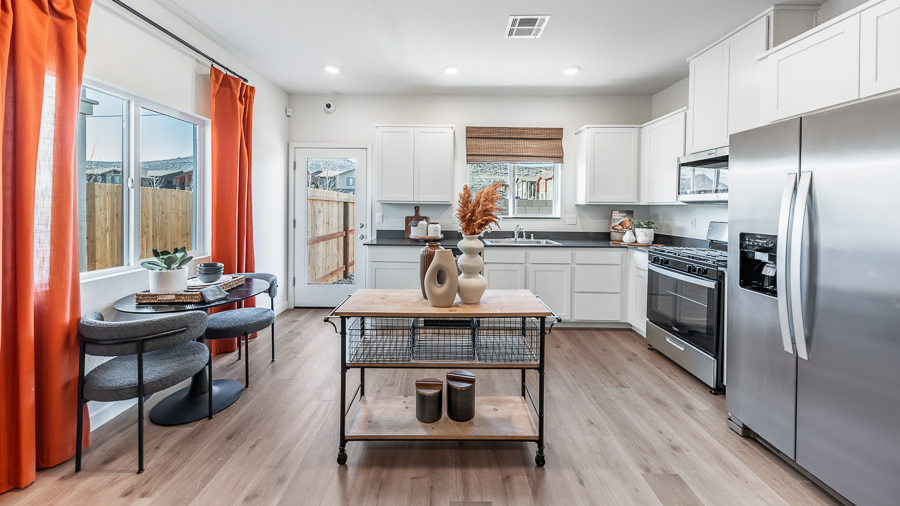 The Denali model home's kitchen boasts white, shaker-style cabinetry, stainless steel applicances, a window over the kitchen sink, a small dining table with two chairs situated beneath a window, and a glass-paned back door.
