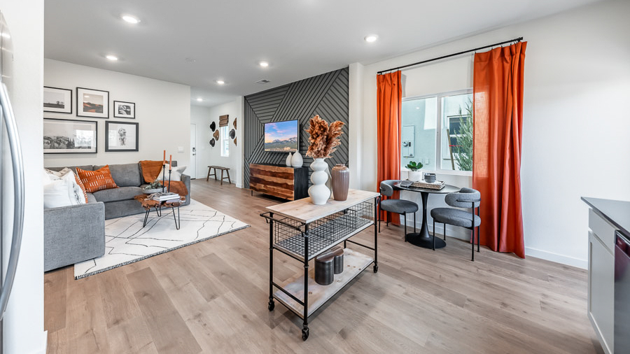 Open living space in Phoenix's model home, with an accent wall with carpentry details behind the wall-mounted TV and console table, a sectional sofa, live-edge coffee table, and a gallery wall of photos behind the couch, a rolling kitchen island, and a cozy dining table with two chairs beneath a window.