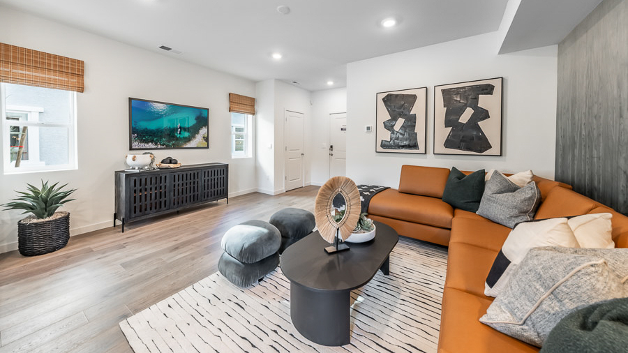A living space in Phoenix's model home with a bold orange sectional sofa, coffee table, and two accent stools, a console table, and a wall-mounted TV flanked by two windows.