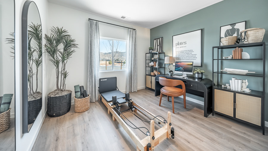 A combination office space and workout room in Phoenix's model home with a desk and desk chair flanked by two bookshelves, a pilates reformer table, a basket with mats and blocks, and two large mirrors mounted on the wall.