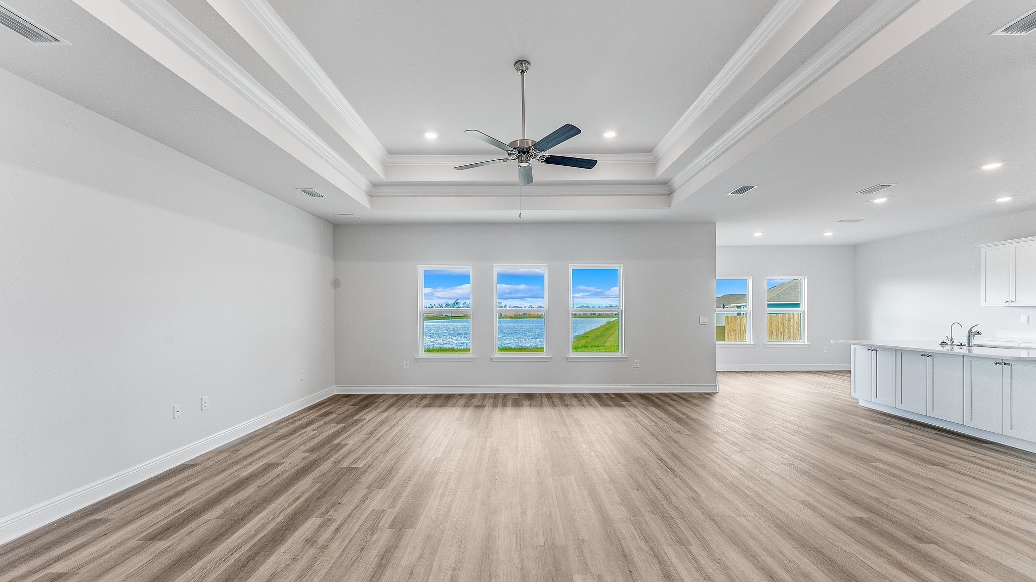 Open living room with EVP flooring and tray ceilings and ceiling fan and 3 windows and kitchen.