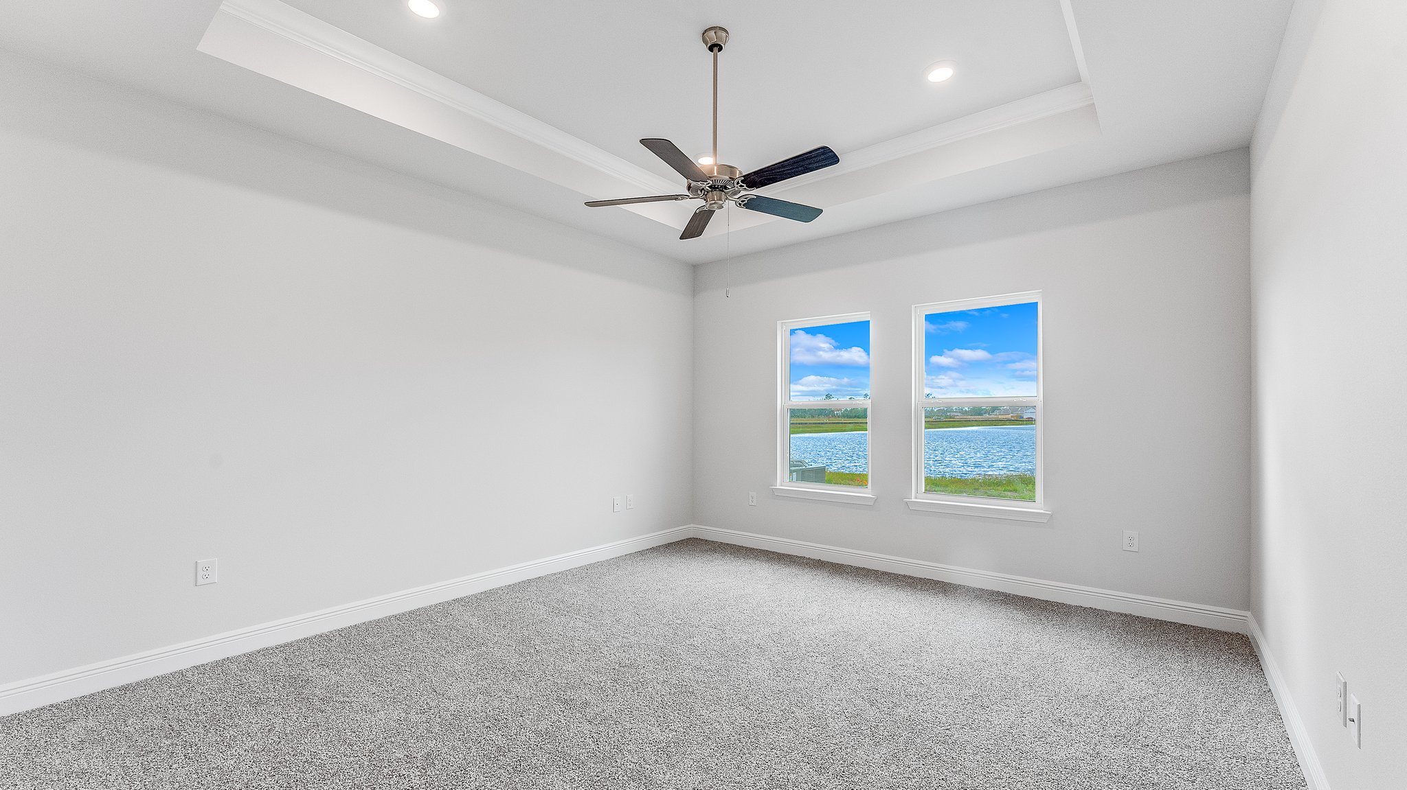 Primary bedroom with carpet floors and ceiling fan and tray ceilings and two windows.