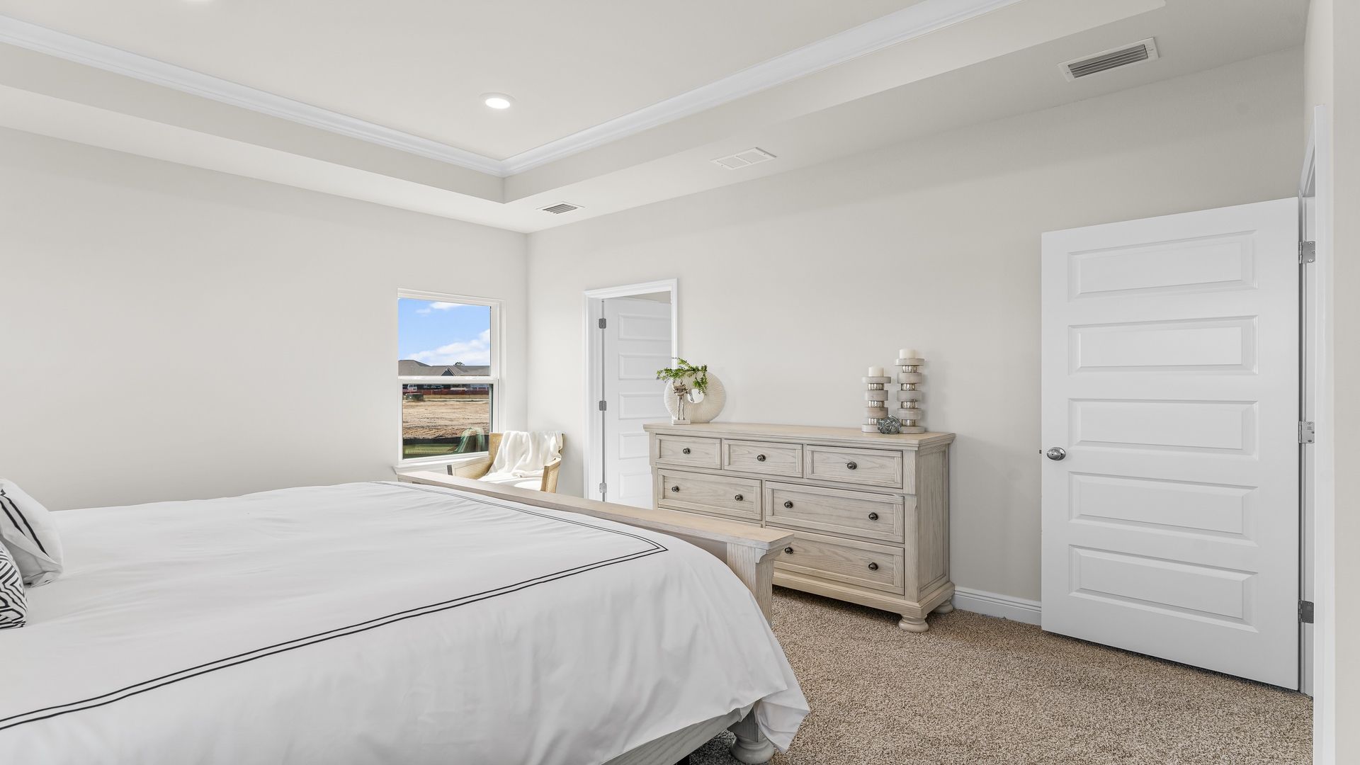 Primary bedroom with carpet floors and tray ceilings with window and bathroom entrance.