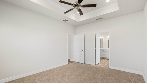 Primary bedroom with carpet floors and tray ceilings and ceiling fan and primary bathroom entrance.
