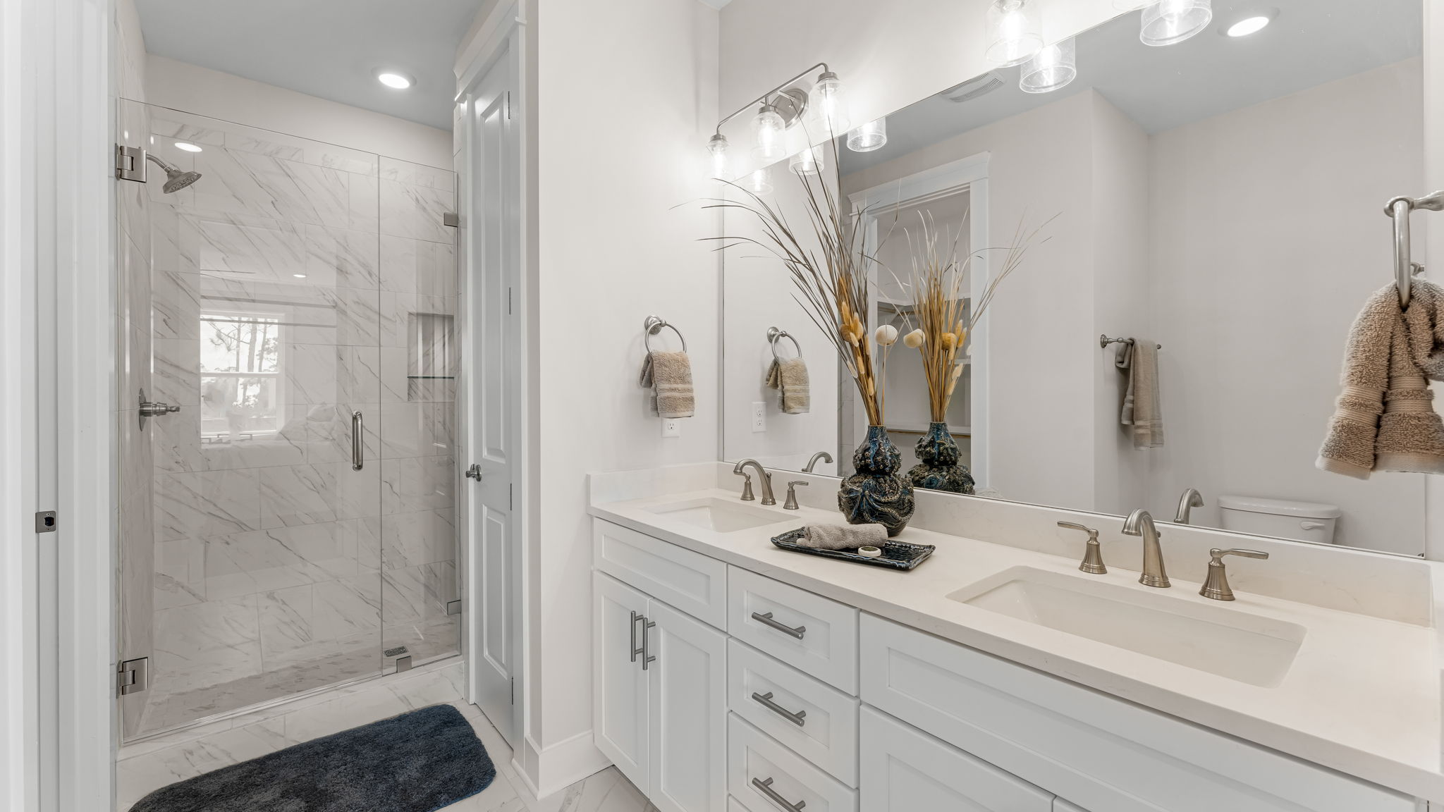 Primary bathroom with double vanity quartz countertops with white cabinets and Florentine tiled shower with frameless glass door.