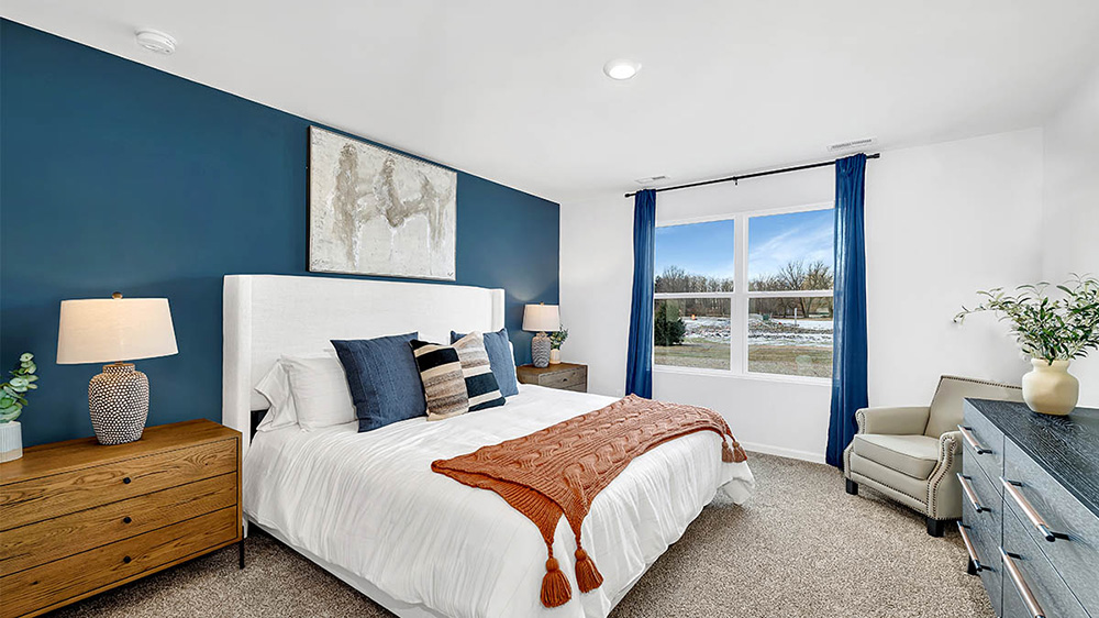 Modern bedroom with a blue accent wall, white bed, and blue pillows. Orange throw adds warmth. Bright window view with blue curtains. Cozy and serene atmosphere.