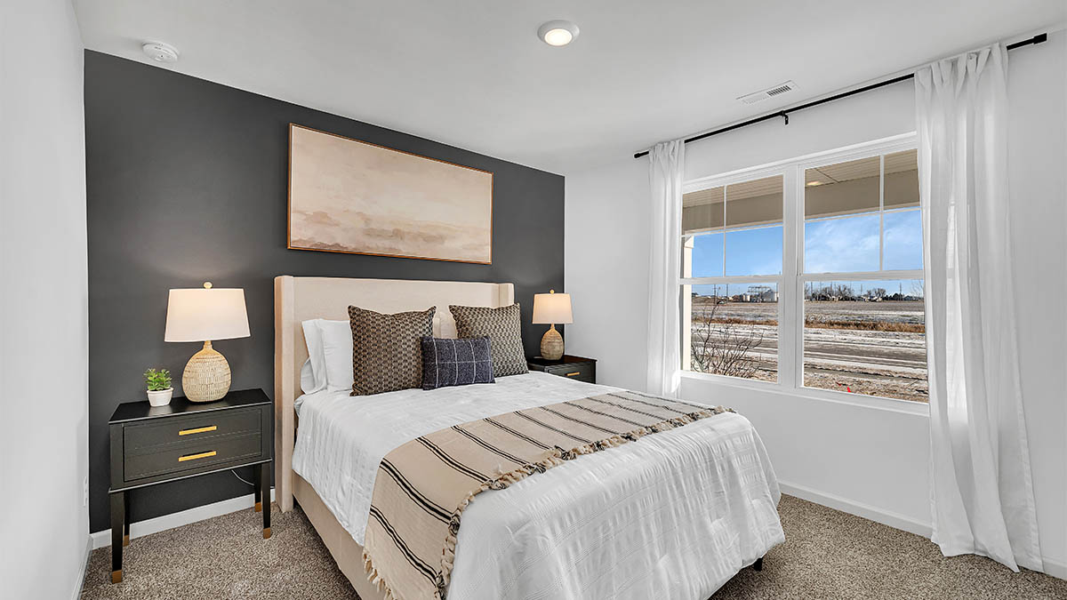 A cozy bedroom with a gray accent wall features a bed with white linens and striped throw, flanked by black nightstands holding lamps. A large window offers a bright, scenic view.