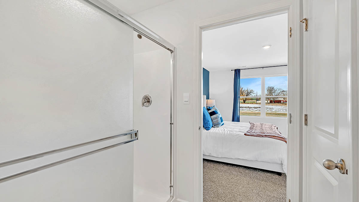 View from a bathroom with a shower into a cozy bedroom. The bedroom features a neatly made bed with blue pillows, blue curtains, and a large window with a snowy landscape outside, creating a serene and inviting atmosphere.