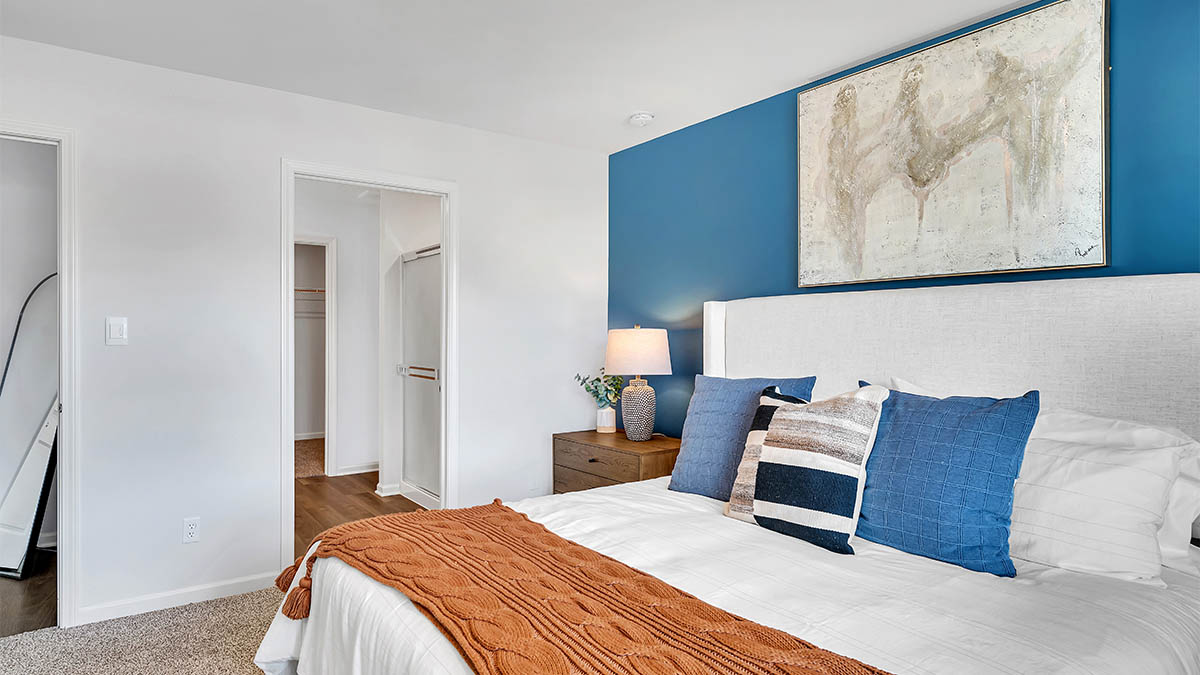 A modern bedroom features a bed with blue and white pillows, a brown throw blanket, and a wooden nightstand with a lamp. A large abstract artwork hangs on a blue accent wall, conveying a calm and stylish atmosphere.