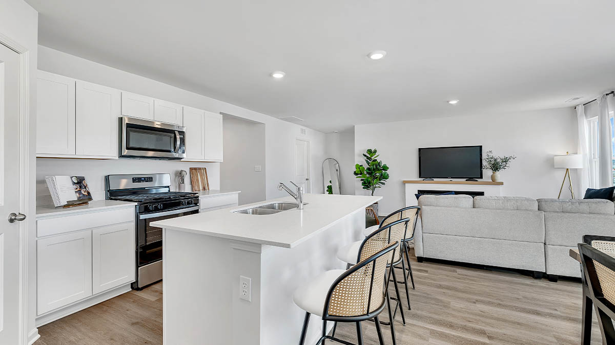 Modern open-concept kitchen and living room. White cabinets, stainless steel appliances, island with bar stools, light wood flooring, neutral decor.