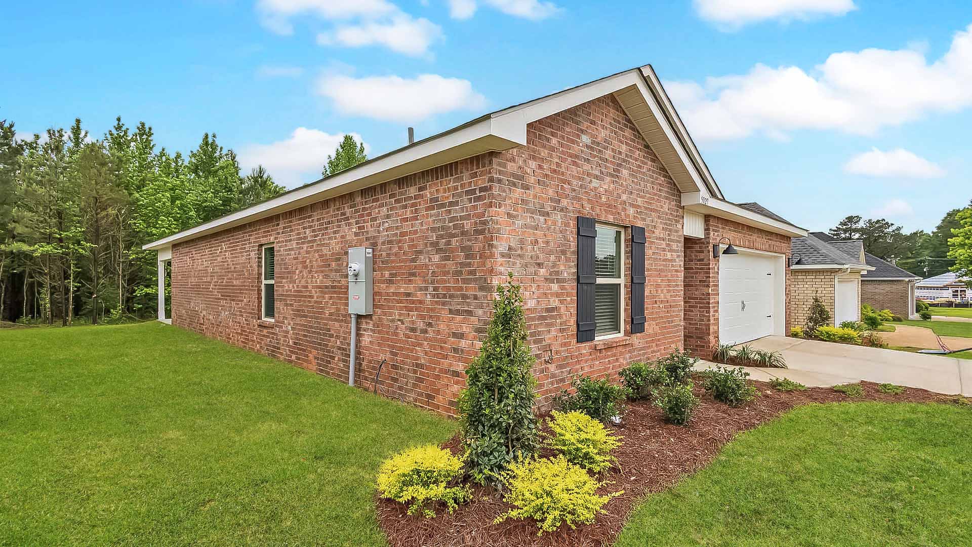 Side exterior single story home with red/brown brick and white paneling and black shutters on the windows with side walk to the front door. The two-car garage and driveway.