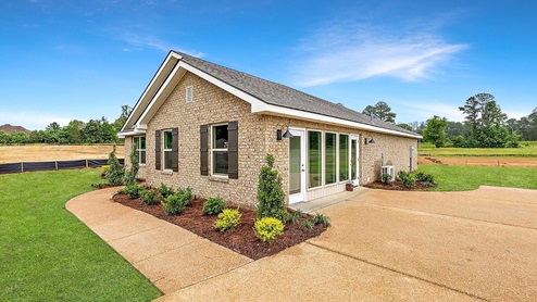 Other side exterior single story home with tan brick and white paneling and black shutters on the windows with side walk to the front door. The two-car garage and driveway are in view.