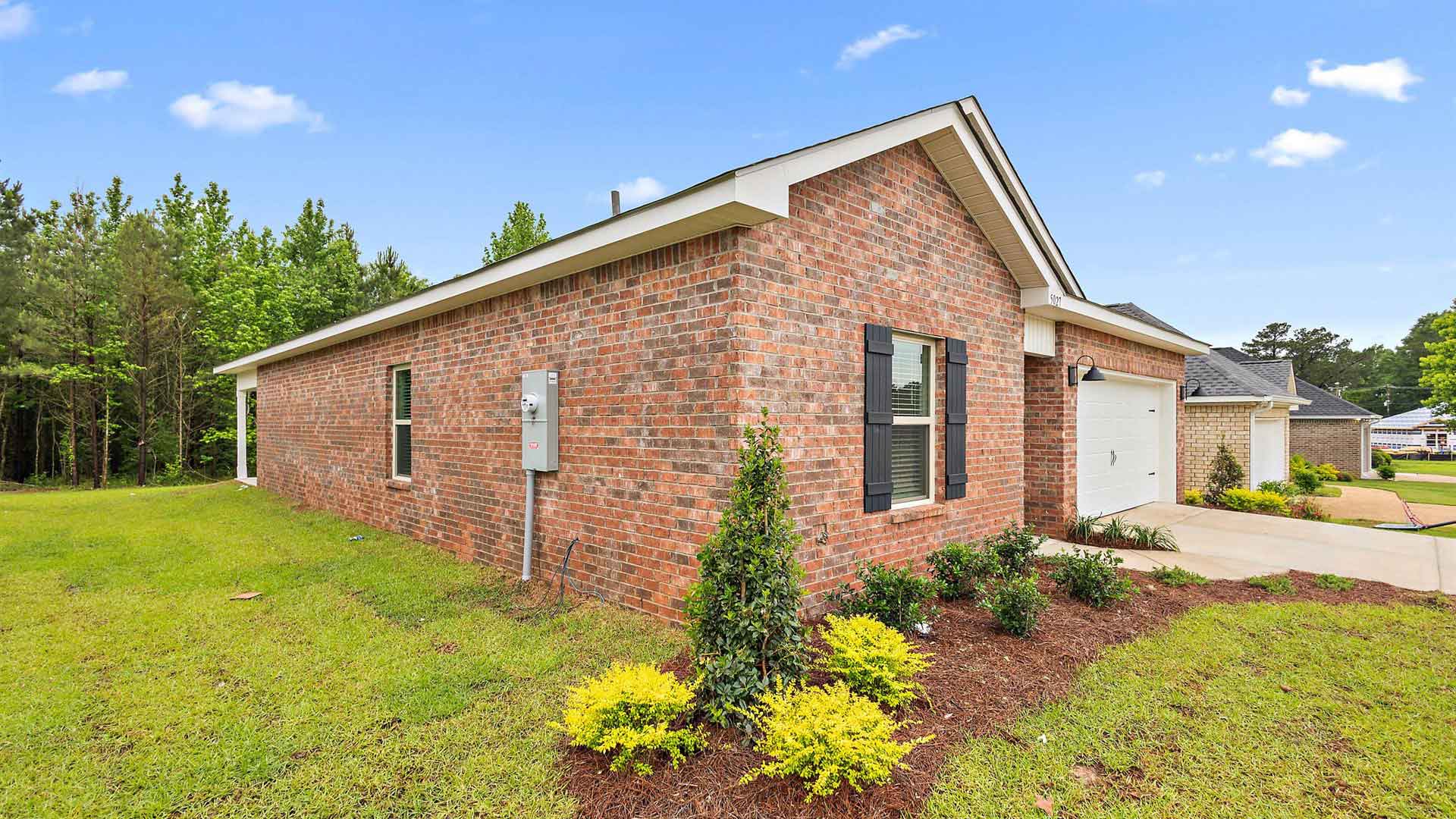 Side exterior single story home with red/brown brick and white paneling and black shutters on the windows with side walk to the front door. The two-car garage and driveway.