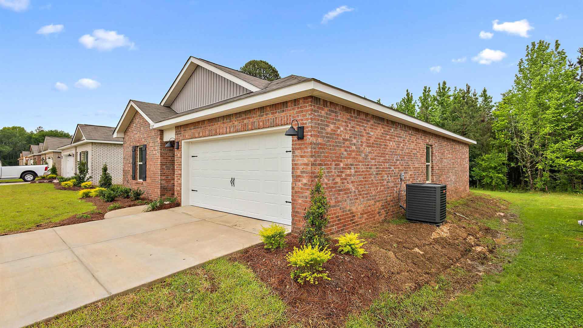 Other side exterior single story home with red/brown brick and white paneling and black shutters on the windows with side walk to the front door. The two-car garage and driveway.