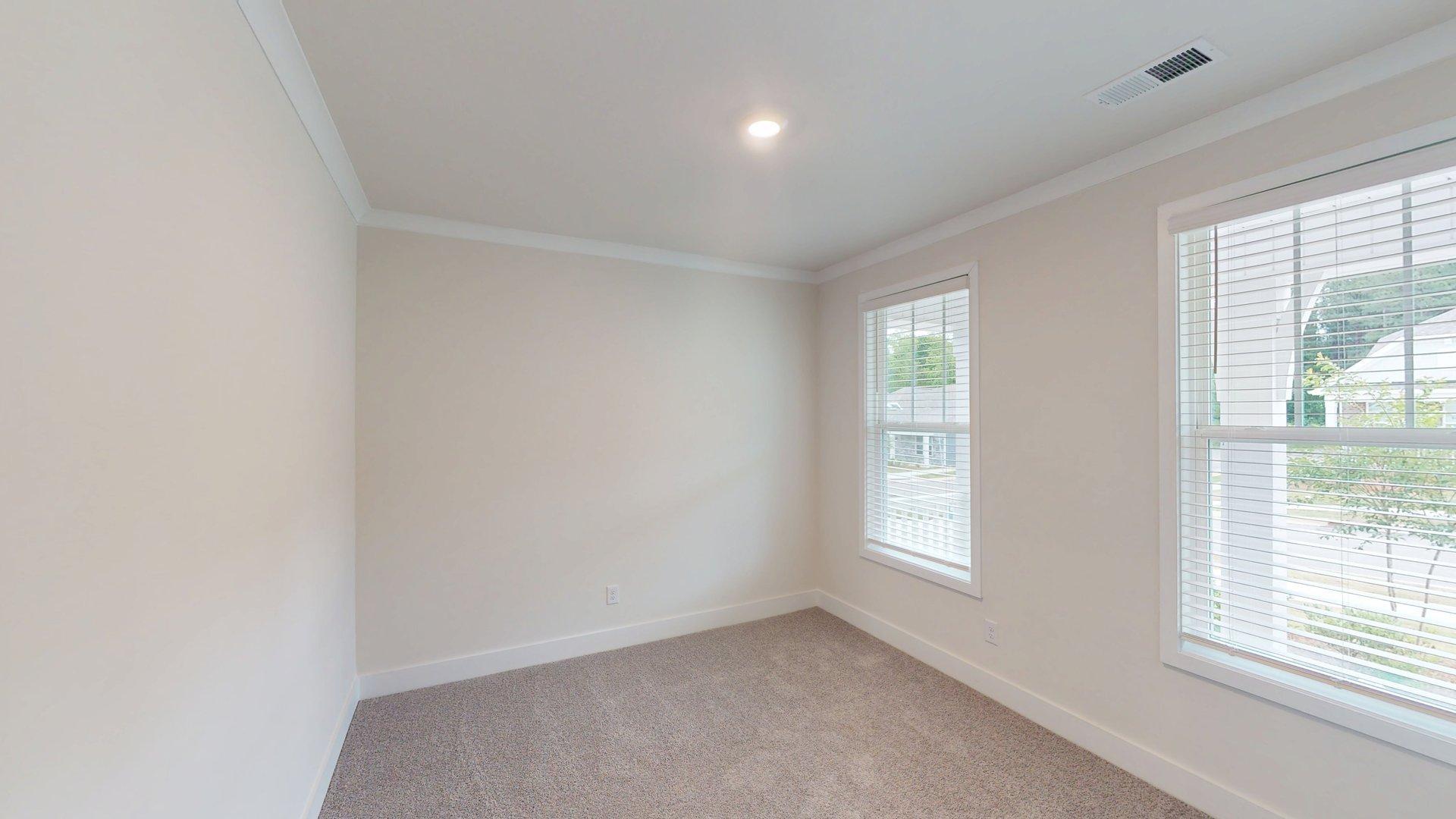 Carpeted bedroom with small window. Primary bathroom with grey cabinets, white countertop, double sink vanity and glass door shower. New Homes in Charlotte, North Carolina