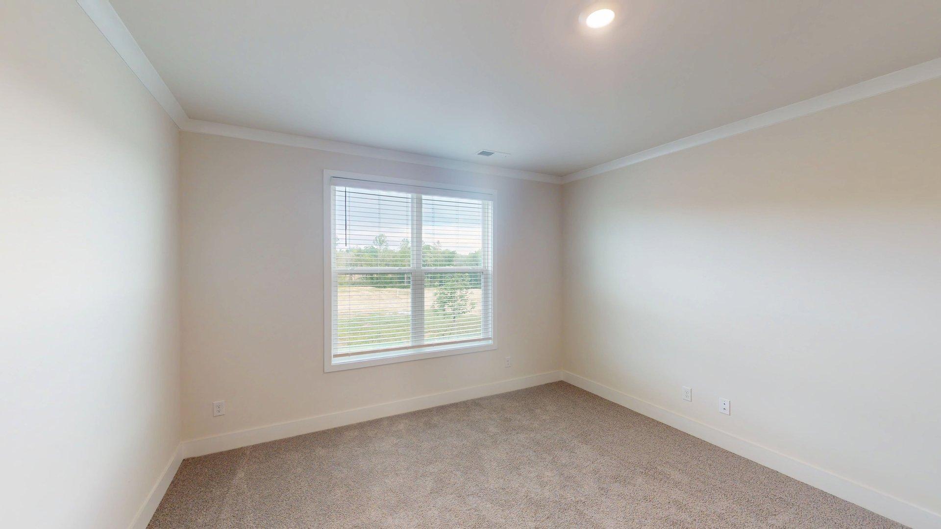 Carpeted bedroom with small window. Primary bathroom with grey cabinets, white countertop, double sink vanity and glass door shower. New Homes in Charlotte, North Carolina