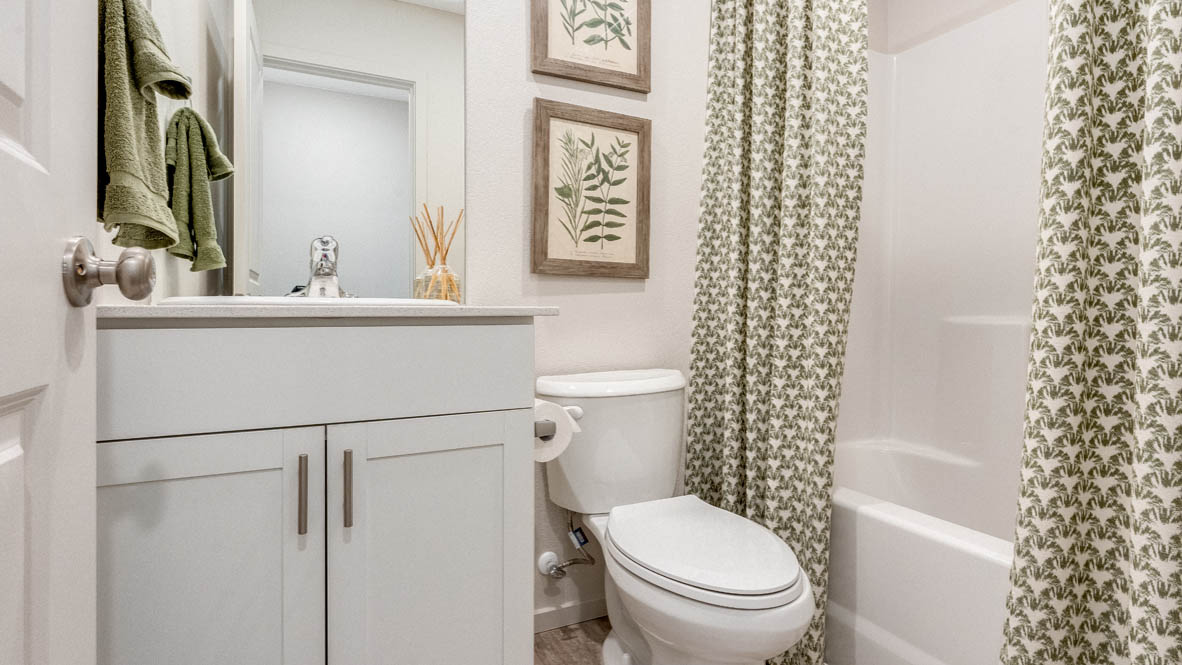East Park Village bathroom with white cabinets, green towels, mirror, quartz countertops, green curtains, two paintings, and a white tub