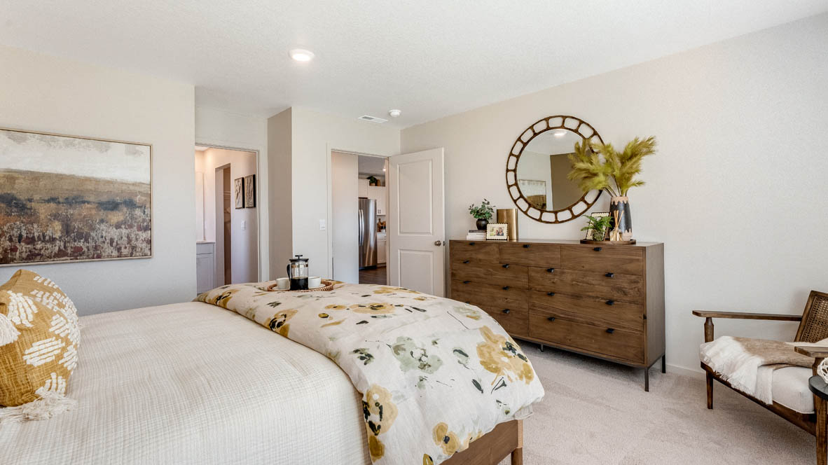 East Park Village Primary bathroom with carpet and a bed with a bedframe, nightstands with lamps, single chair with paintings, windows, and dresser with mirror