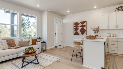 Gordon Creek Pointe Great Room with a coffee table, white couch with pillows, windows, and the kitchen with island peninsula that has quartz countertops, and stools