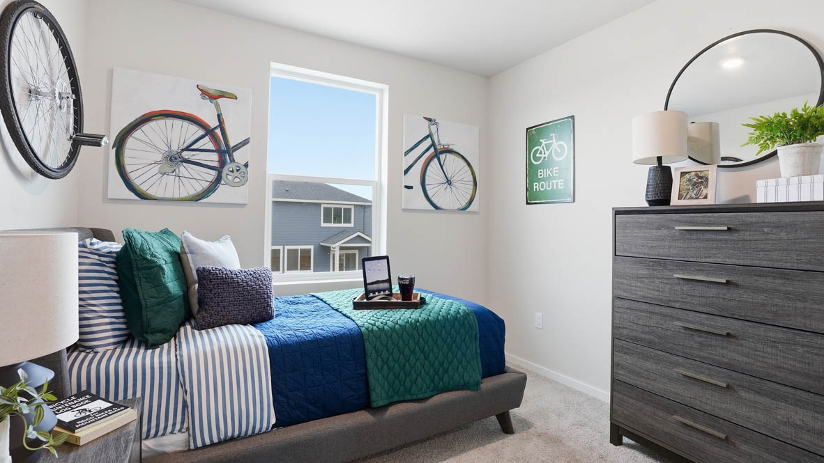 Stevens Ranch Townhomes bedroom with a gray dresser, two lamps, circle mirror, bike paintings, bed with a blue comforter, teal accent blanket, accent pillows, striped sheets, carpeting, and a window