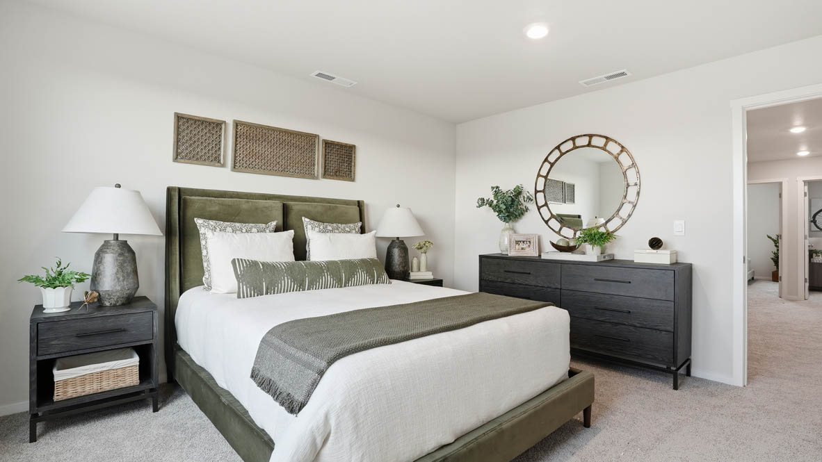 Stevens Ranch Townhomes primary bedroom with two windows, carpeting, white walls, olive bedding with white sheets, black nightstands, two lamps, circle mirror, and a dresser