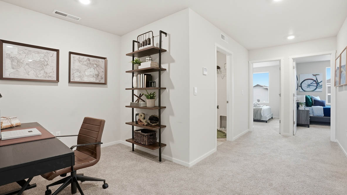Stevens Ranch Townhomes loft with a black office desk, brown rolling desk chair, shelving with decor, two hung up world maps, carpeting, white walls, the bedrooms in the background