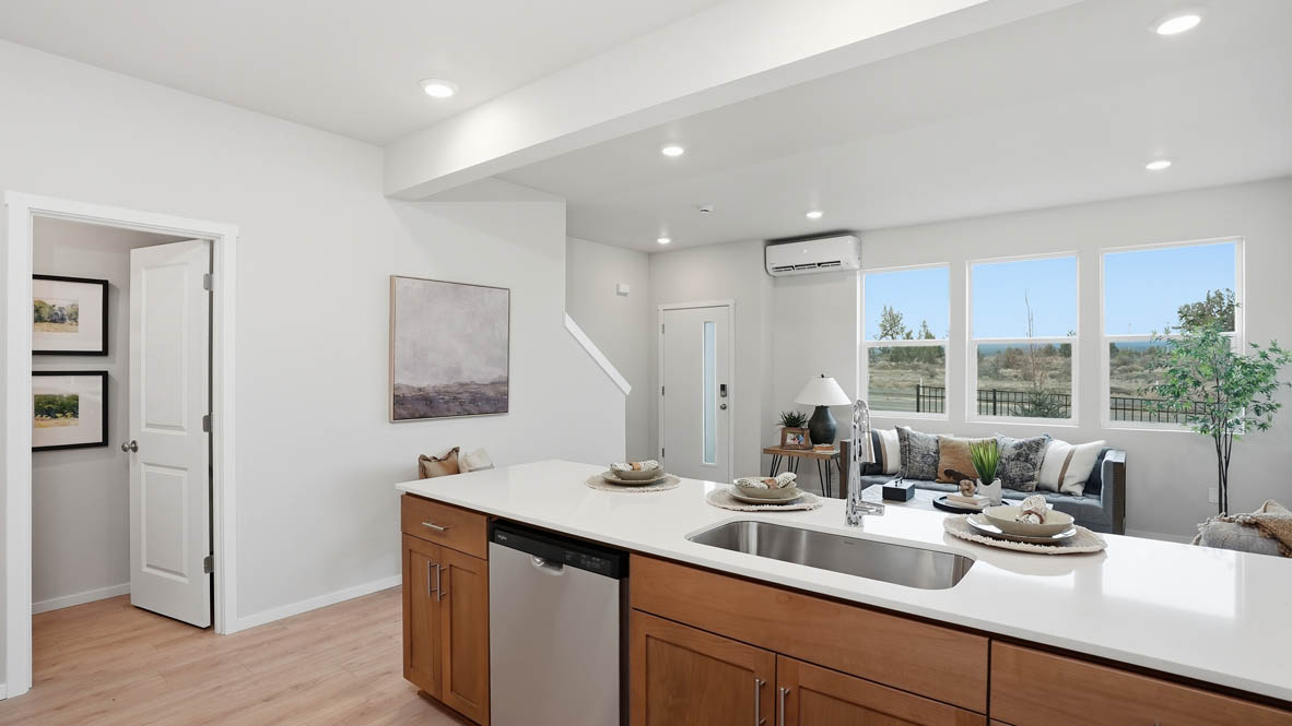 Stevens Ranch Townhomes kitchen with stained soft-closed cabinets, solid surface countertops, decorative plate sets, three stools, stainless steel appliances, laminate flooring, and the great room in the background