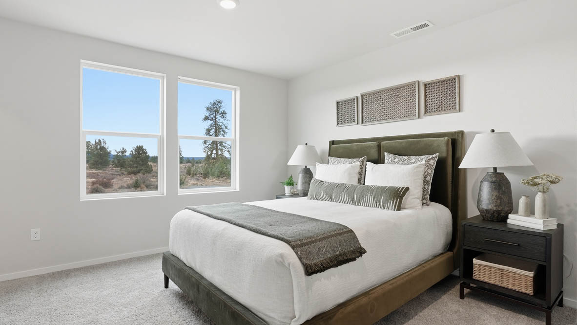 Stevens Ranch Townhomes primary bedroom with two windows, carpeting, white walls, olive bedding with white sheets, black nightstands, two lamps, circle mirror, and a dresser