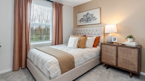 Lowrie Estates bedroom with light beige accent wall, white bedding, orange window drapes, and wooden accent table