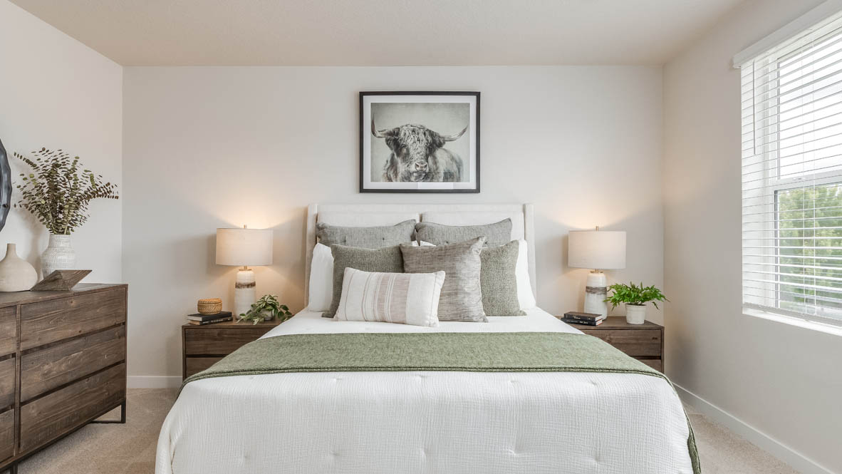 Davis Estates Primary Bedroom with white comforter and a green accent blanket, pillows, two nightstands with windows, center panting, dresser with a plant on top, and window