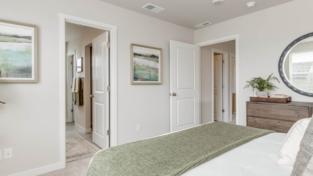 Davis Estates Primary bedroom with white comforter and green accent blanket, dresser with a plant, circle mirror, two paintings, and view of the primary bathroom