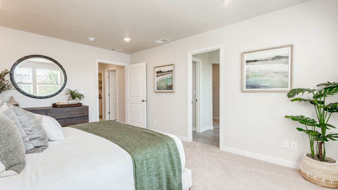 Davis Estates Primary bedroom with white comforter and green accent blanket, dresser with a plant, circle mirror, two paintings, and view of the primary bathroom