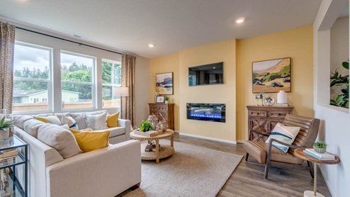 Marcola Meadows great room with a yellow accent wall with a fire place and a tv mounted above it, an l shaped sectional and coffee table over an area rug, with a brown chair and two brown wooden accent cabinets on either side of the tv
