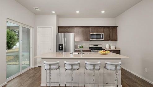A kitchen with brown wood cabinets and stainless steel appliances, there's a large island in the middle with a quartz countertop and four white stools. There's glass sliding doors to the left leading to the backyard.