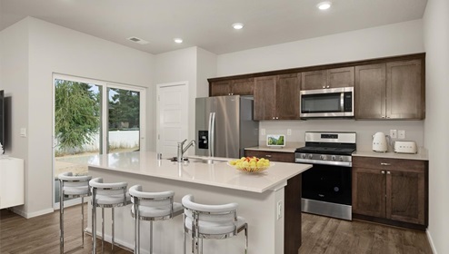 A kitchen with brown wood cabinets and stainless steel appliances, there's a large island in the middle with a quartz countertop and four white stools. There's glass sliding doors to the left leading to the backyard.