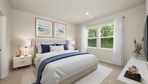 A bedroom with a large bed with white sheets and decorative pillows and two night stands featuring lamps. There are two pictures hanging from the wall and two large windows letting in natural light framed by sheer white curtains.