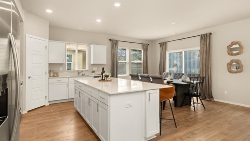 A kitchen with quartz countertops and white cabinets, a refridgerator to the left, and three burnt orange stools in front of the kitchen island. There's a window on the far wall letting in natural light.