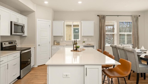 A kitchen with quartz countertops and white cabinets, a refridgerator to the left, and three burnt orange stools in front of the kitchen island. There's a window on the far wall letting in natural light.