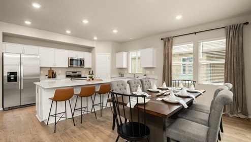 A dining area with a wooden table set with napkins and plates and off white dining chairs with a view of both the kitchen with white cabinets and a fridge and the living area with two abstract art pieces hanging above the couch.