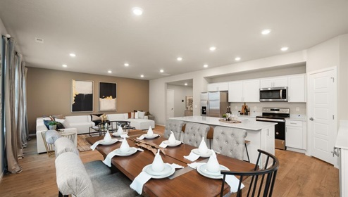 A dining area with a wooden table set with napkins and plates and off white dining chairs with a view of both the kitchen with white cabinets and a fridge and the living area with two abstract art pieces hanging above the couch.
