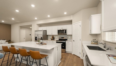 A kitchen with quartz countertops and white cabinets, a refridgerator to the left, and three burnt orange stools in front of the kitchen island. There's a window letting in natural light.