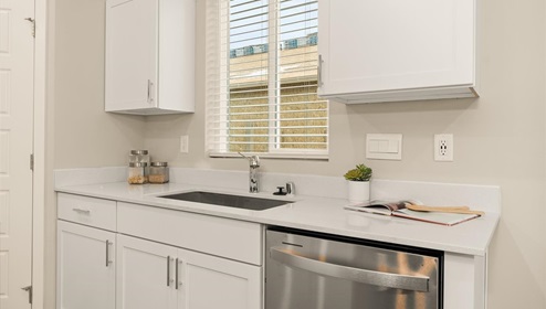 A kitchen with quartz countertops and white cabinets, a refridgerator to the left, and three burnt orange stools in front of the kitchen island. There's a window letting in natural light.