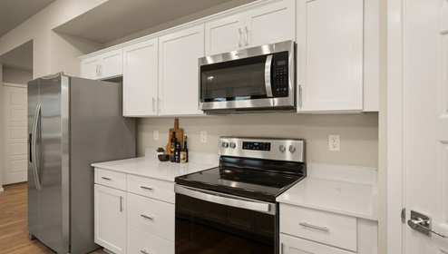 A kitchen with quartz countertops and white cabinets, a refridgerator to the left, and three burnt orange stools in front of the kitchen island. There's a window letting in natural light.