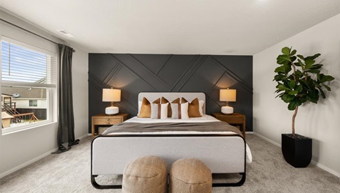 A large bedroom with a dark grey accent wall with a large bed with white and grey bedding and decorative pillows. There's two nightstands featuring lamps and a dresser against the far wall with an art piece hung above. There's three large windows letting in natural light framed by dark curtains.