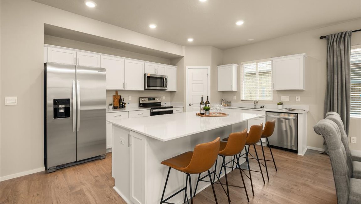 A dining area with a wooden table set with napkins and plates and off white dining chairs with a view of both the kitchen with white cabinets and a fridge and the living area with two abstract art pieces hanging above the couch.