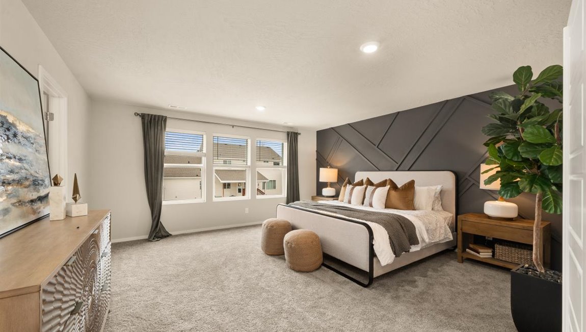 A large bedroom with a dark grey accent wall with a large bed with white and grey bedding and decorative pillows. There's two nightstands featuring lamps and a dresser against the far wall with an art piece hung above. There's three large windows letting in natural light framed by dark curtains.