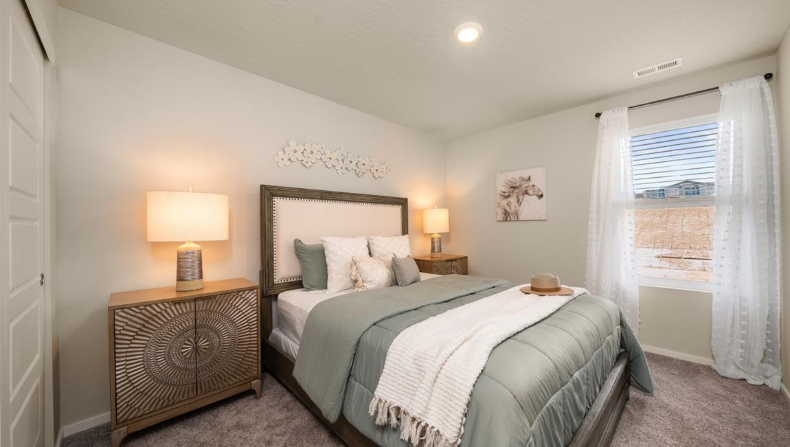 A bedroom featuring a bed with muted green sheets and decorative pillows. There are two nightstands with lamps and a window letting in natural light framed by sheer white curtains.