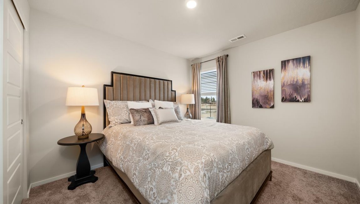 A bedroom featuring a bed with beige floral patterned sheets and decorative pillows. There are two nightstands with lamps and abstract art on the right wall next to a window letting in natural light framed by grey curtains.