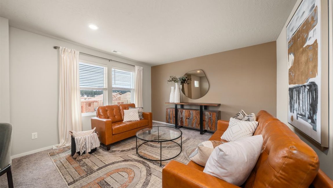 A living area with a large patterned ornamental rug with burnt orange couches and a glass coffee table sitting on top. There's a console sitting against the far wall and abstract art hanging on the right wall. There are two large windows letting in natural light framed by sheer white curtains.